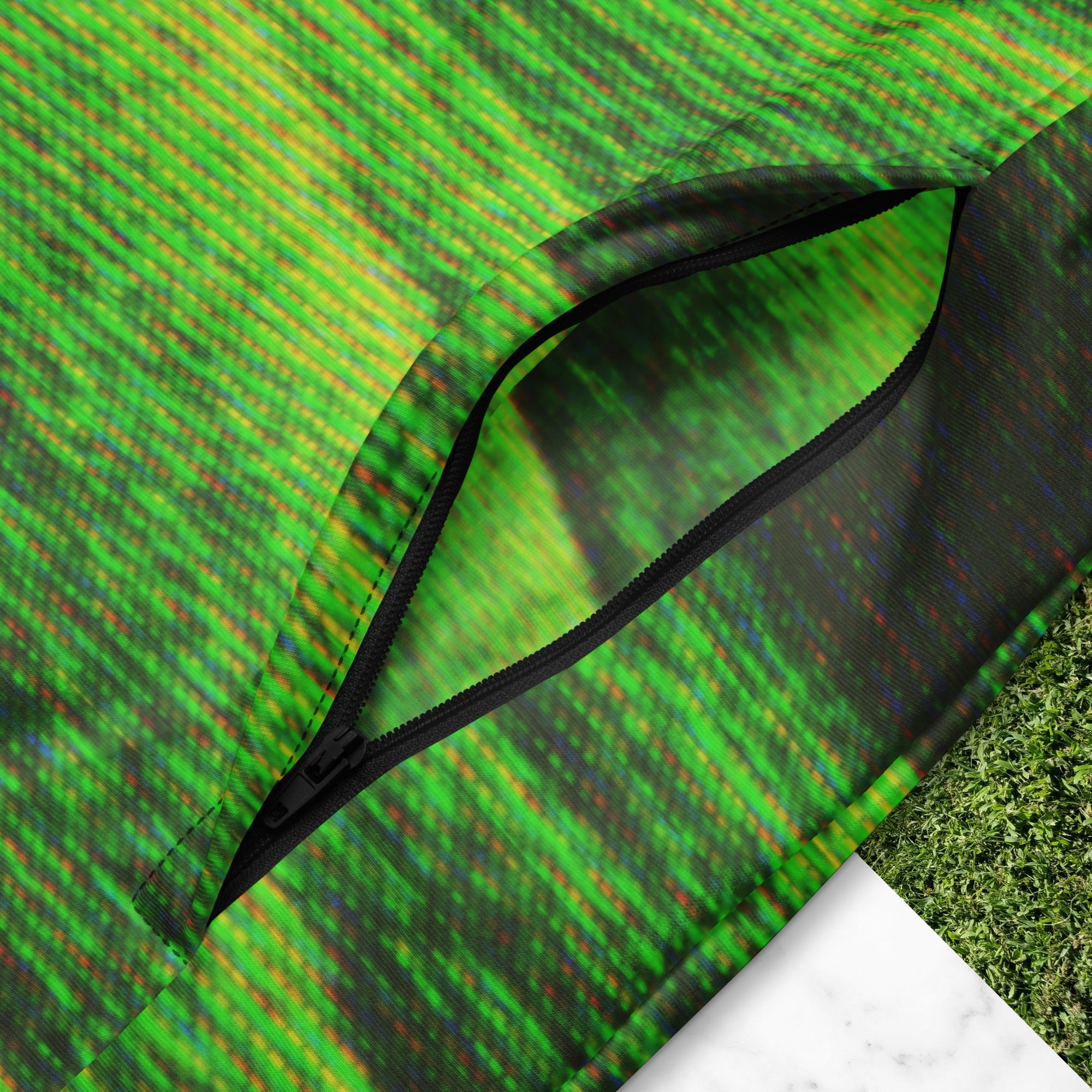 Closeup of pocket zipper on primarily green and black full color all over print track jacket depicting grass covered green rolling hills and their shadows overlayed with detailed v h s and retro t v distortion with a green and blue Neon Pools vaporwave sun and waves emblem on the left chest