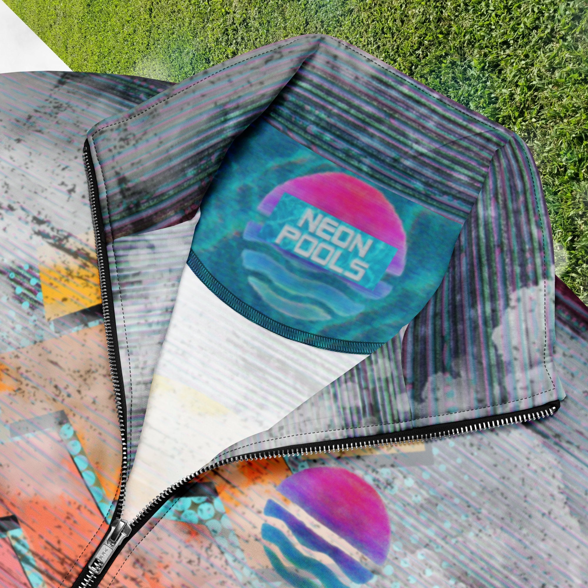 Closeup of neckline and Neon Pools inner logo and insignia on colorful vaporwave and memphis design inspired all over print track jacket depicting a foreground of grass and palm trees with a backdrop of mountains more mountains and a sky filled with retro neon orange to yellow cascading bold zigzags and a vaporwave broken sun on the back with lots of clouds various bars and geometric shapes on the sleeves with Neon Pools magenta and blue vaporwave sun and waves logo at left chest